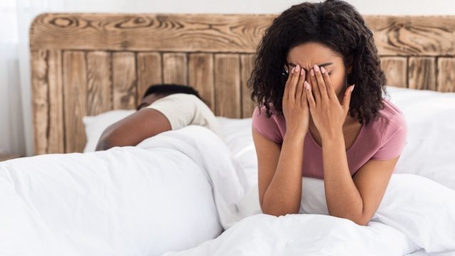 woman sitting up in bed rubbing her eyes while male partner sleeps