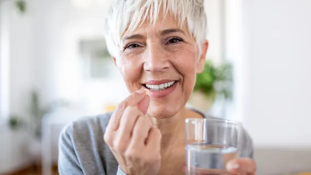 A happy mature woman with short white hair wearing a gray long-sleeved shirt takes a vitamin with a glass of water