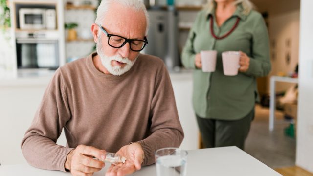 A senior man sitting at a table and taking vitamins or medicine while someone brings him a drink