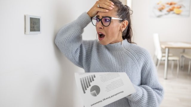 upset woman holding a heating bill and looking at her thermostat